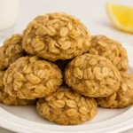 A plate of Lemon Oatmeal No-Bake Cookies stacked in a pyramid shape, with visible oats and a golden hue, accompanied by a lemon wedge and glass of milk in the background.